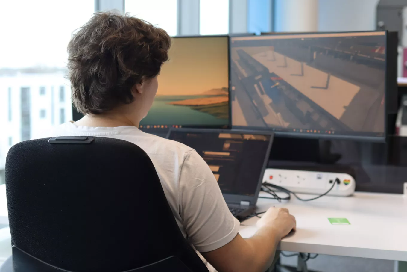 An engineering designer at Equans Sci-Tech working at a triple-monitor station, meticulously reviewing a detailed 3D architectural render and facility layout.